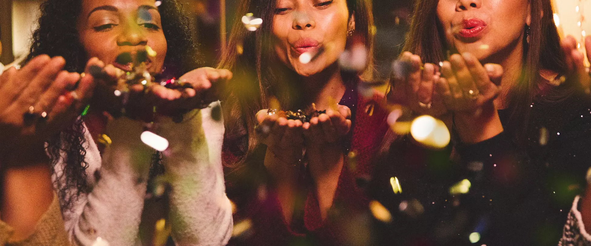 Three women facing the camera. Their eyes are closed and their hands are raised to their mouths. They are about to blow gold confetti from their hands..