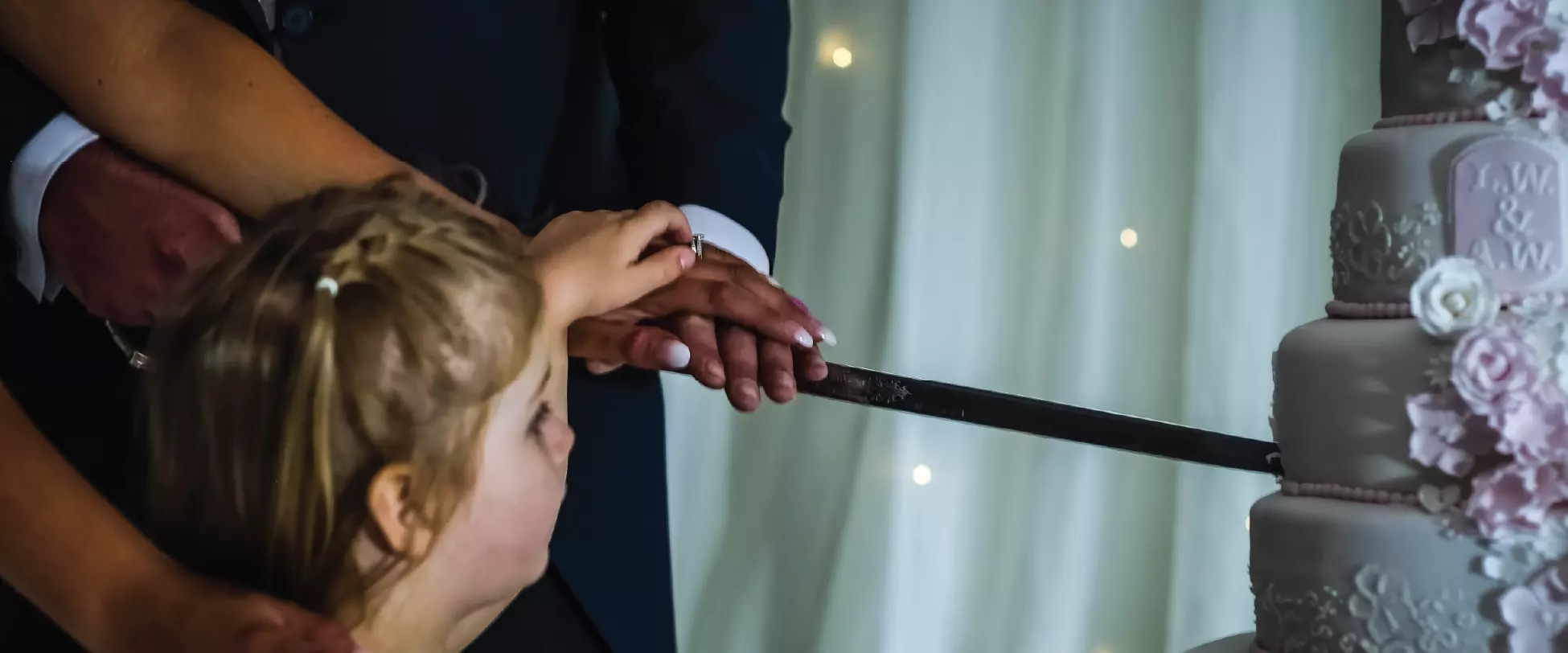 A bride and grooms hands are holding a knife to cut their wedding cake. A young girl in frame is helping them..